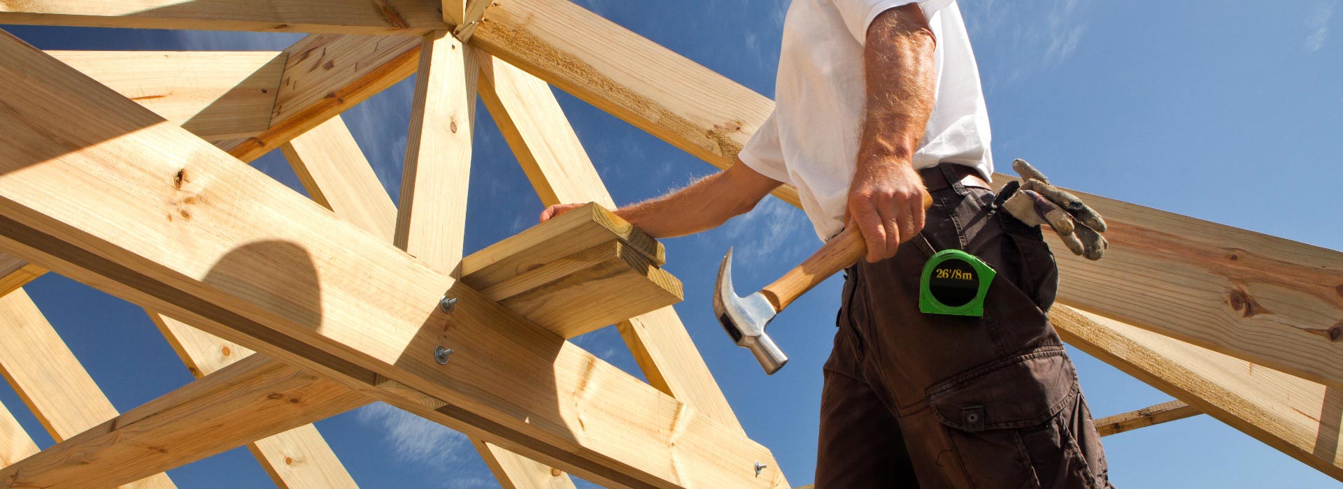 Person hält Hammer, trägt weißes Hemd, steht auf einem Holzbalken, blauer Himmel im Hintergrund.