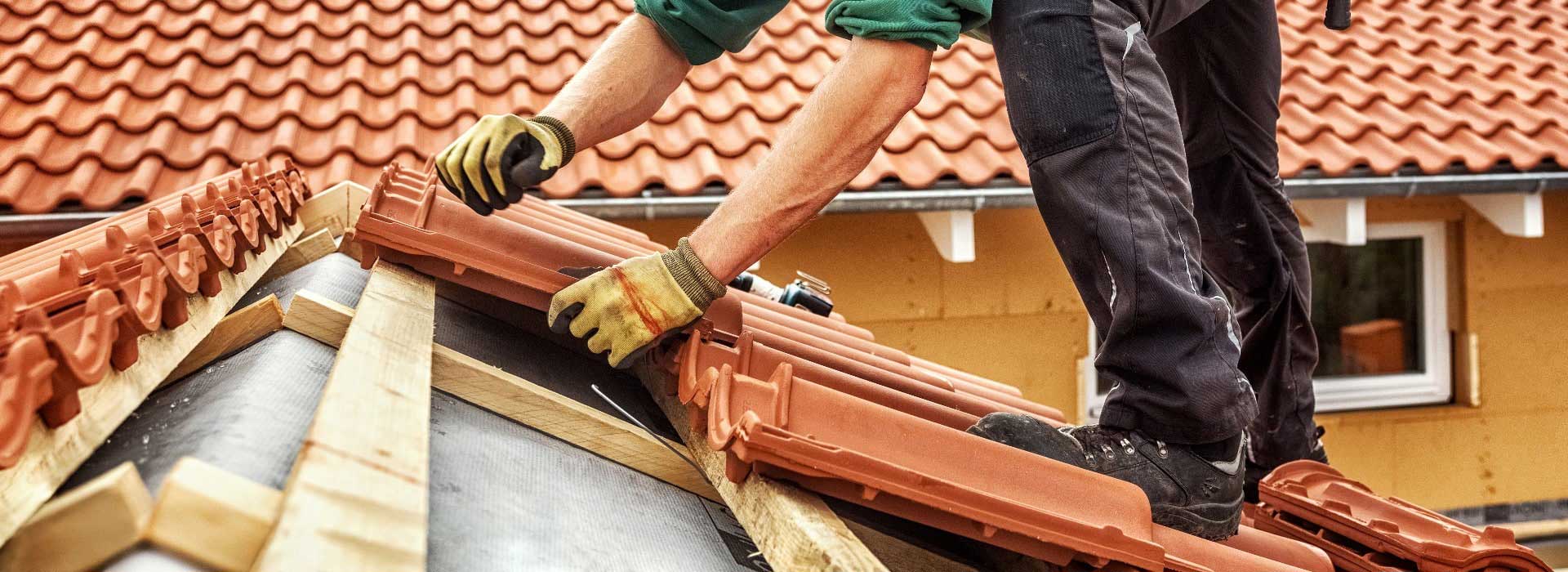 Handwerker deckt Dach mit roten Ziegeln, Fokus auf Hände und Werkzeug, trägt Handschuhe und Arbeitskleidung.